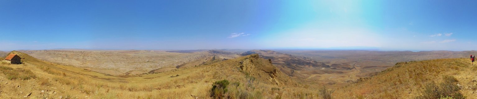 Wide panoramic photograph of dry golden-brown hills and valleys under a clear blue sky near Signagi, Georgia, with a small building on the left and two hikers visible on the right ridge
