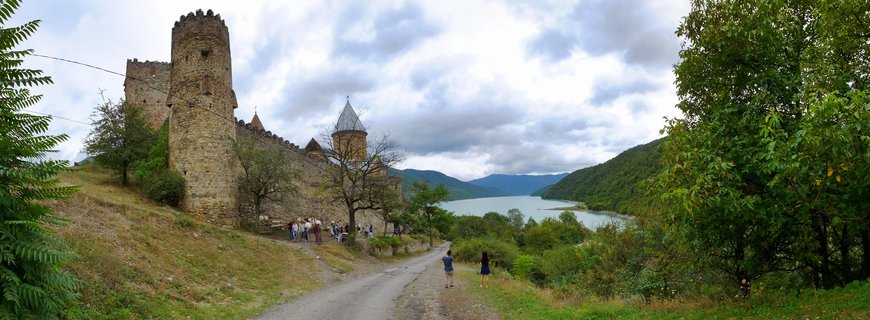 Medieval stone fortress with round towers and conical church roofs on a green hillside overlooking a blue reservoir with mountains in the background under cloudy skies