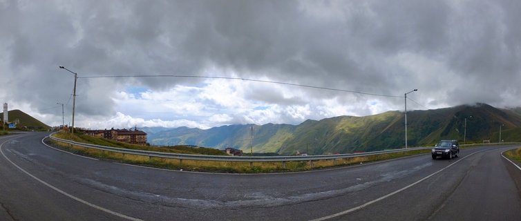 Panoramic view of a winding asphalt road through green Caucasus mountains under cloudy skies with a vehicle and roadside buildings visible