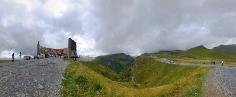 Panoramic view of the Russia-Georgia Friendship Monument stone structure with colorful murals on a mountainside overlooking green Caucasus mountain valleys under cloudy sky with tourists on gravel viewing platforms