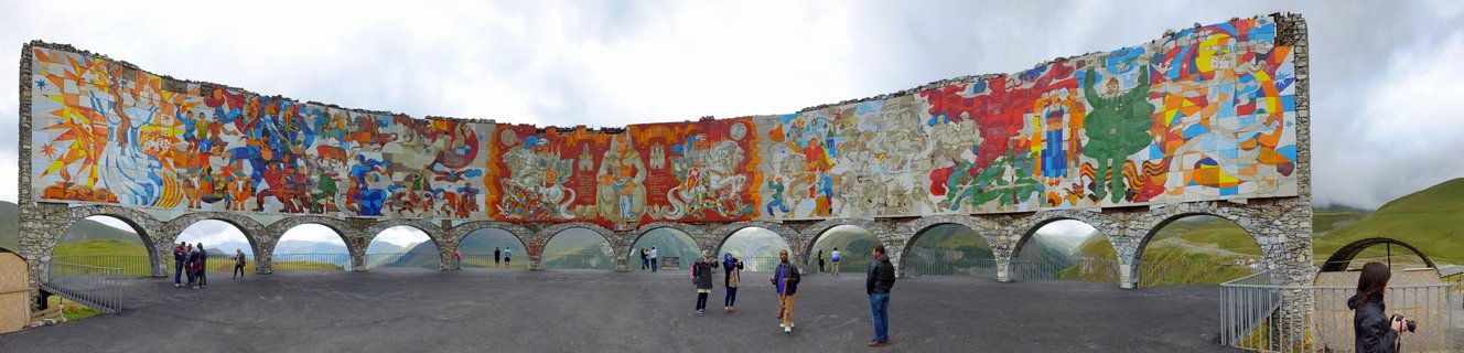 A curved stone monument wall covered in colorful mosaic murals with multiple arches below, tourists walking on paved plaza, overcast sky and mountains visible in background