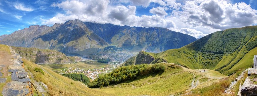 Panoramic mountain landscape showing a valley town surrounded by green hills with dramatic rocky mountain peaks and white clouds in blue sky, Kazbegi region, Georgia