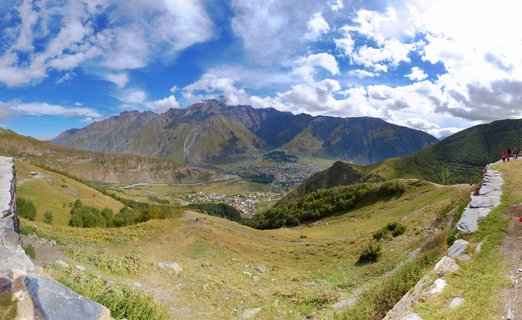 Wide mountain valley landscape showing green hills with stone walls in foreground, a town in the valley center, and large rocky mountains in the background under blue sky with white clouds