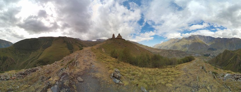 Panoramic view of Gergeti Trinity Church monastery on a grassy hilltop in the Caucasus Mountains with dramatic cloudy sky and mountain ranges in the background