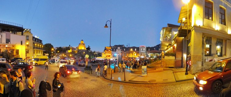 Wide panoramic view of a busy cobblestone street in Tbilisi old town at twilight with pedestrians, cars, illuminated buildings, and a church visible on a hill in the background