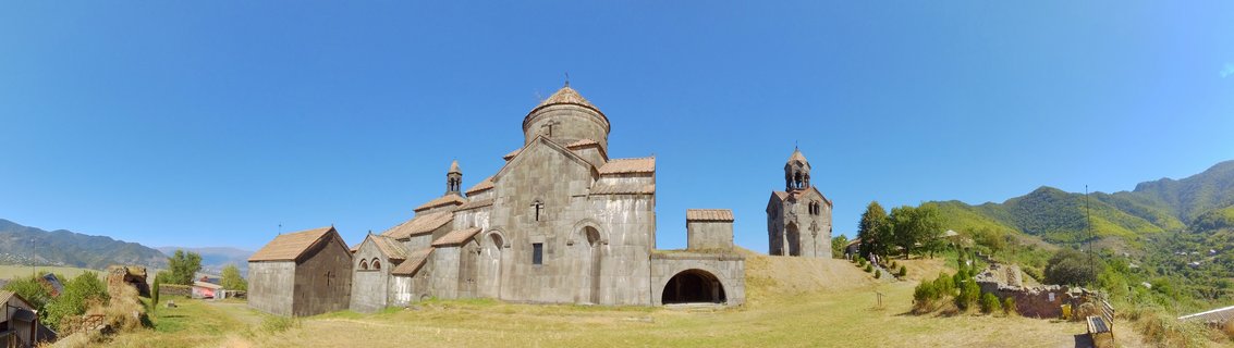 Medieval Armenian monastery complex with stone churches, conical domes, and bell tower on grassy hillside with mountains in background under clear blue sky