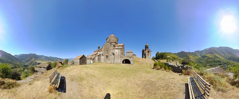 Medieval Armenian stone monastery with conical dome and bell tower on grassy hillside, mountains and blue sky background