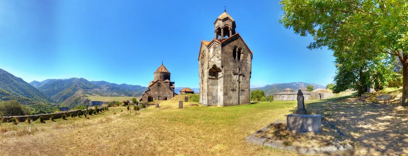 Panoramic view of medieval stone monastery buildings with conical domes on grassy hillside with mountains and blue sky in background
