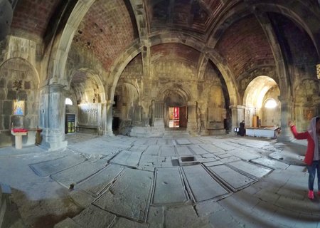 Interior of historic Armenian monastery showing stone arches, vaulted ceilings, ancient tomb slabs on floor, religious icons on walls, and visitors exploring the sacred space