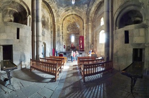 Interior view of a stone church with arched ceilings, wooden pews, a large chandelier, and several visitors standing near the altar area with red religious banners
