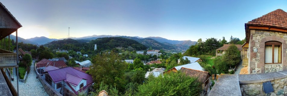 Panoramic view of traditional stone houses with red-tiled roofs in Dilijan, Armenia, surrounded by green trees and forested mountains under a clear blue sky at sunset