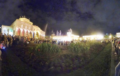 Night photograph of Republic Square in Yerevan showing illuminated buildings, lit fountains, and crowds of people gathered in the plaza with landscaped gardens in the foreground.