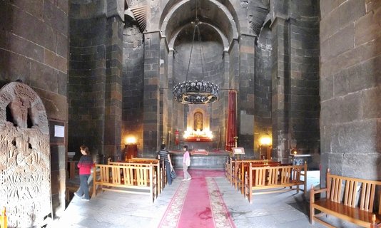 Church interior with dark stone walls, arched ceiling, hanging chandelier, red carpet aisle, wooden benches, illuminated altar with religious icon, and carved stone cross on left wall