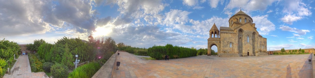 Stone Armenian church with central dome and bell tower on paved courtyard under cloudy blue sky with sun visible