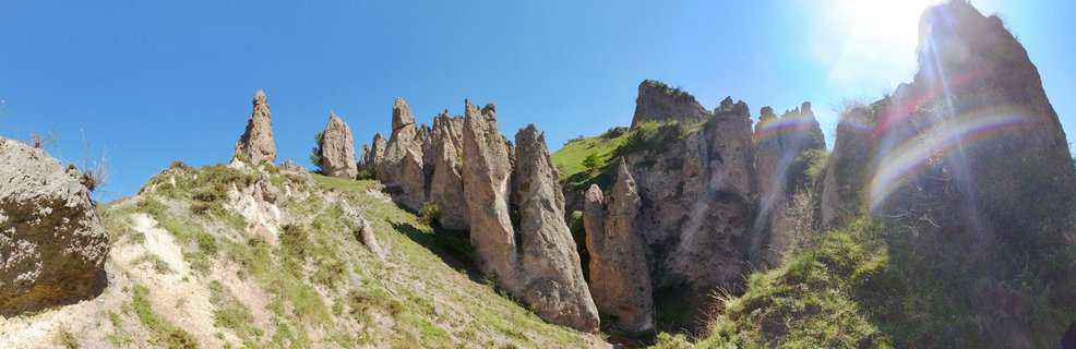 Tall natural rock spires and pinnacles on a green grassy hillside under bright blue sky with sun flare
