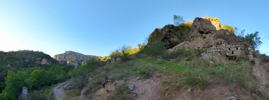 Ancient stone cave dwellings carved into a rocky hillside with green vegetation, dirt path, and clear blue sky in Goris, Armenia
