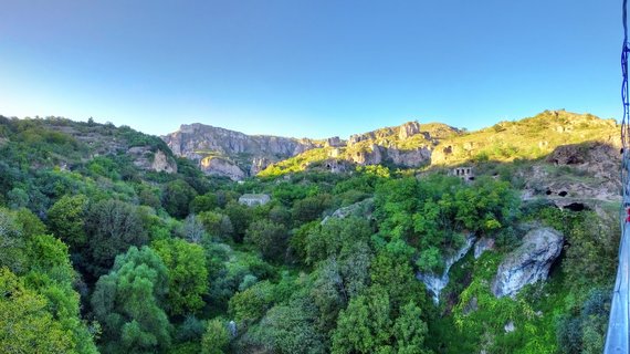 Panoramic view of ancient cave dwellings carved into rocky cliffs above a green forested valley in Goris, Armenia under clear blue sky