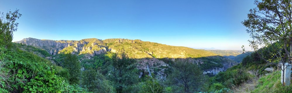 Wide panoramic landscape showing green mountain valley with rocky limestone cliffs and ancient cave dwellings under clear blue sky in Goris, Armenia