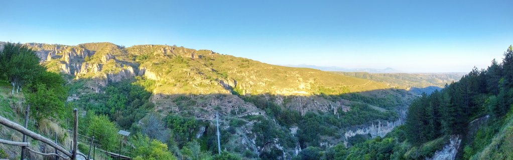 Panoramic landscape showing a green mountain valley in Goris, Armenia with rocky cliffs, cave dwellings carved into hillsides, trees, and a wooden fence in the foreground under a clear blue sky