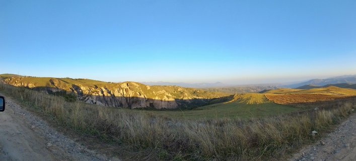 Wide panoramic landscape showing rolling green and golden hills with rocky cliff formations under clear blue sky in Goris, Armenia