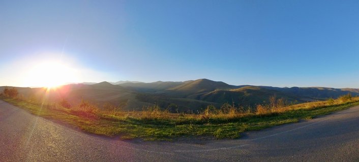 Panoramic landscape showing rolling green and brown mountains under a clear blue sky with the sun setting on the left, casting warm light over hills with a curved asphalt road in the foreground