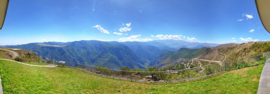 Panoramic mountain valley landscape showing a deep gorge with forested slopes, metal safety railing at viewpoint, winding access roads, and distant blue mountain ranges under clear sky
