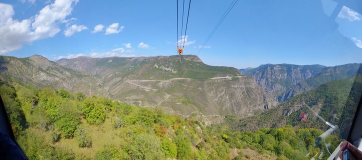 View from inside a cable car showing an orange gondola suspended on cables over a deep mountain valley with green slopes, winding roads, and blue sky with scattered clouds