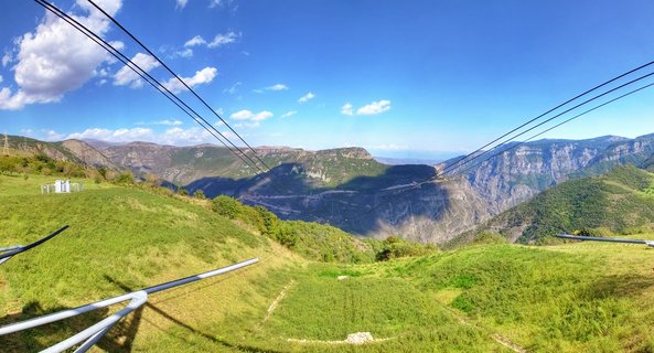 Panoramic view from Wings of Tatev cable station showing four parallel steel cables across Vorotan River Gorge. Vibrant green grassy hillsides slope toward deep mountain canyon with rocky cliffs. Azure blue sky with scattered white cumulus clouds. Silver metal railings in foreground.