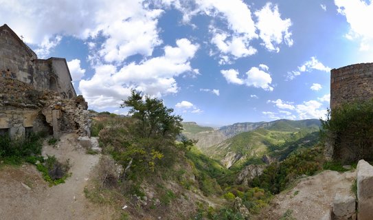 Ancient stone monastery buildings on a clifftop edge overlooking a deep green canyon valley with mountain ranges in the distance under blue sky with white clouds