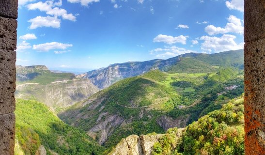 Mountain valley landscape viewed through ancient stone columns showing green hills, deep canyon, winding roads, and blue sky with white clouds in Tatev, Armenia