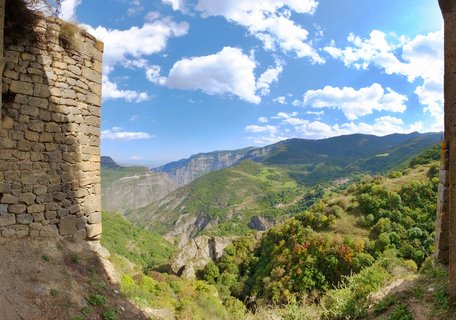 Mountain valley landscape viewed through ancient stone walls, showing green forested slopes, distant mountain ranges, and blue sky with white clouds in Armenia