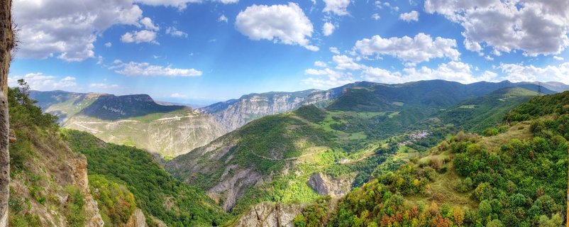 Wide panoramic landscape showing green forested mountains and rocky cliffs surrounding a deep valley gorge in Armenia with blue sky and white clouds above