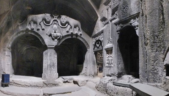 Stone interior of Armenian cave monastery with carved arches, lion reliefs above doorways, and religious iconography on volcanic rock walls