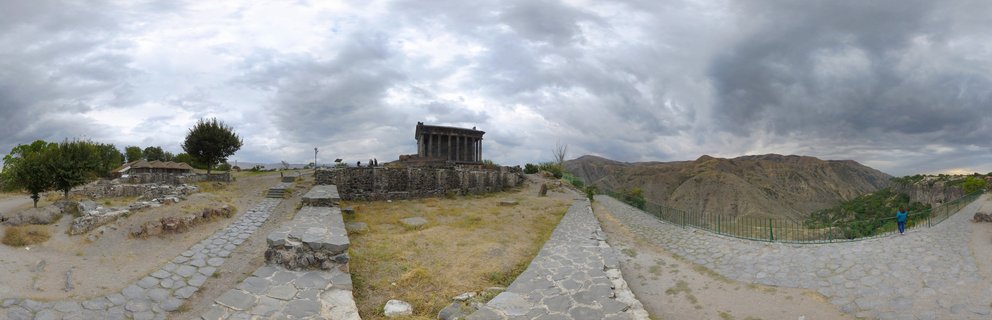 Ancient stone temple with columns surrounded by archaeological ruins, stone pathways, dry grass, mountains in background under cloudy overcast sky