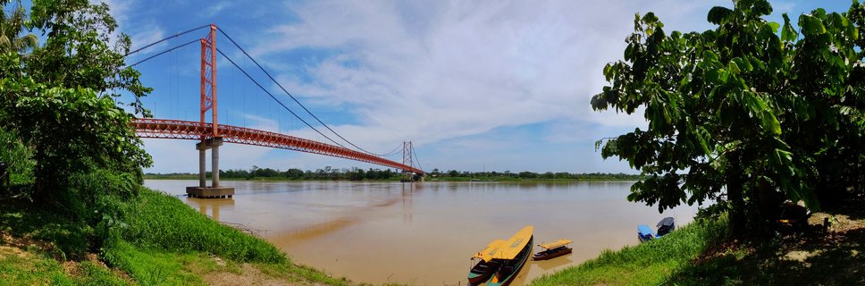 Orange suspension bridge spanning wide brown river with small boats on grassy bank, tropical trees framing the scene under blue sky with clouds in Puerto Maldonado, Peru