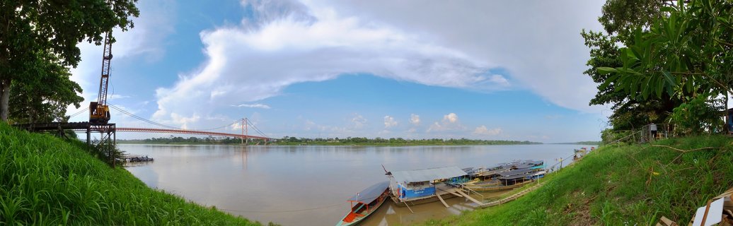 Wide river view showing a red suspension bridge spanning muddy brown water, with docked boats along a grassy riverbank, tropical trees on both sides, and blue sky with white clouds above