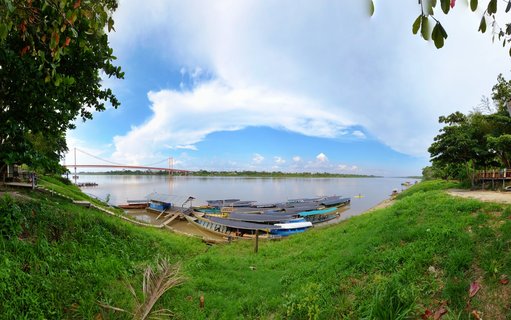 Wide river view showing multiple covered boats docked on grassy banks with a red suspension bridge spanning the water and tropical trees framing the scene under blue sky with clouds