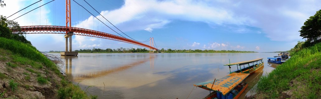 Orange suspension bridge spanning wide muddy river with traditional boats docked at riverbank, lush green vegetation on both shores under blue sky with white clouds in Puerto Maldonado, Peru