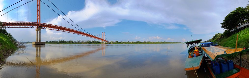 Orange suspension bridge spanning wide brown river with boats docked at riverbank, green rainforest vegetation on both shores under blue sky with clouds in Puerto Maldonado, Peru