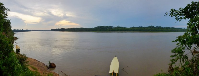 Wide river view showing two white-covered boats moored at muddy riverbank with dense tropical rainforest on opposite shore under cloudy sky