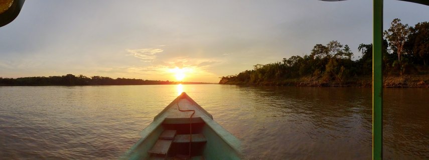 View from the bow of a wooden boat traveling on a wide brown river toward a setting sun, with dense green tropical forest lining both shores