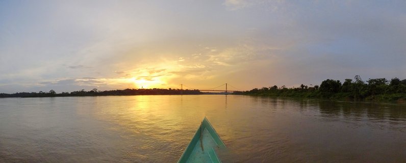 Wide panoramic river view at sunset showing teal boat bow in foreground, golden sunlight reflecting on muddy brown water, suspension bridge in distance, dense green rainforest on both banks, and dramatic cloudy sky with warm orange and yellow tones