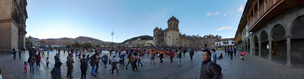 Wide view of Plaza de Armas in Cusco showing the cathedral, colonial buildings with arches, cobblestone plaza filled with tourists, and mountains in the distance under blue sky