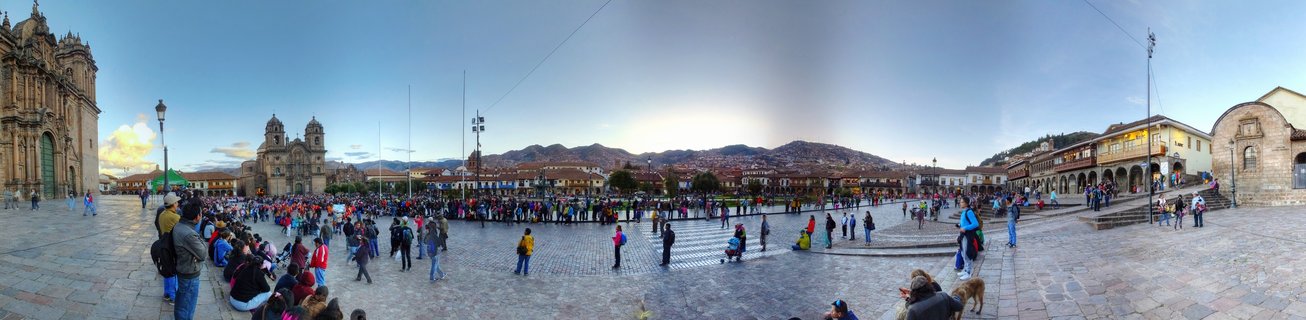 Wide panoramic view of Plaza de Armas in Cusco showing colonial cathedral, stone plaza with crowds of people, colonial buildings with arcades, and Andes mountains in the background under partly cloudy sky
