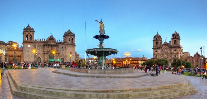 Panoramic view of Plaza de Armas in Cuzco, Peru at dusk, featuring a central bronze fountain with Pachacutec statue, the colonial baroque Cuzco Cathedral with twin bell towers on the left, and the Church of the Society of Jesus on the right, with tourists walking across the stone plaza under illuminated street lamps against a clear blue evening sky