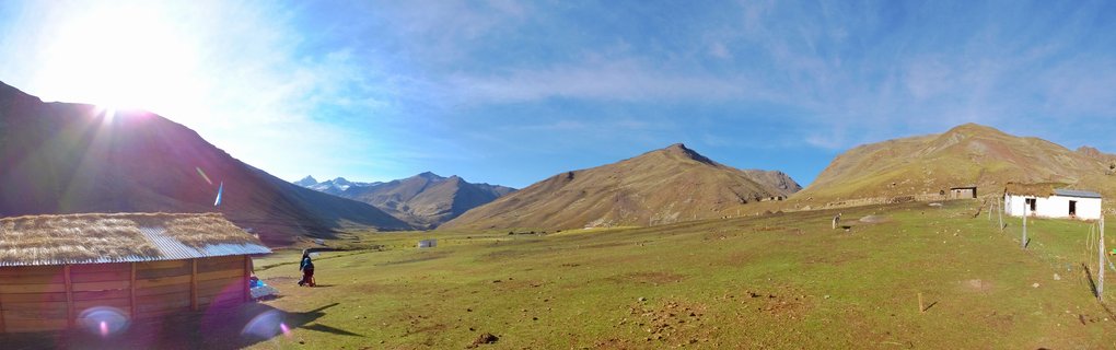 Wide landscape showing Andean mountain valley with wooden buildings, green-brown grassland, distant snow-capped peaks, and bright sun in blue sky