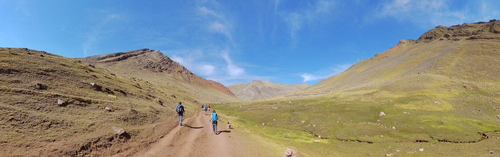 Group of hikers walking on a dirt trail through a mountain valley with colorful stratified mountains in the background under a clear blue sky in Peru
