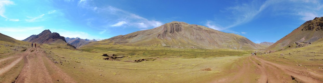 Panoramic view of mountainous valley in Peru with dirt hiking paths, small figures of people walking, green and brown grasslands, and large striped Rainbow Mountain under blue sky with wispy clouds