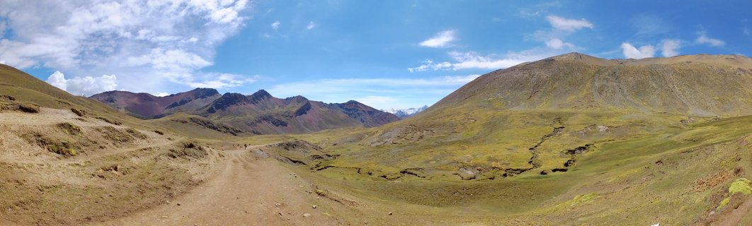 Wide valley landscape in Peru with dirt hiking trail leading toward colorful striped Rainbow Mountain peaks under blue sky with white clouds