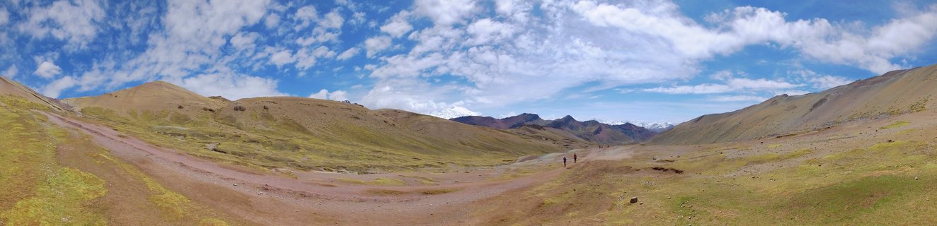 Panoramic landscape showing a valley with rolling hills in brown and green tones, a dirt path with two hikers, distant snow-capped mountains, and blue sky with white clouds